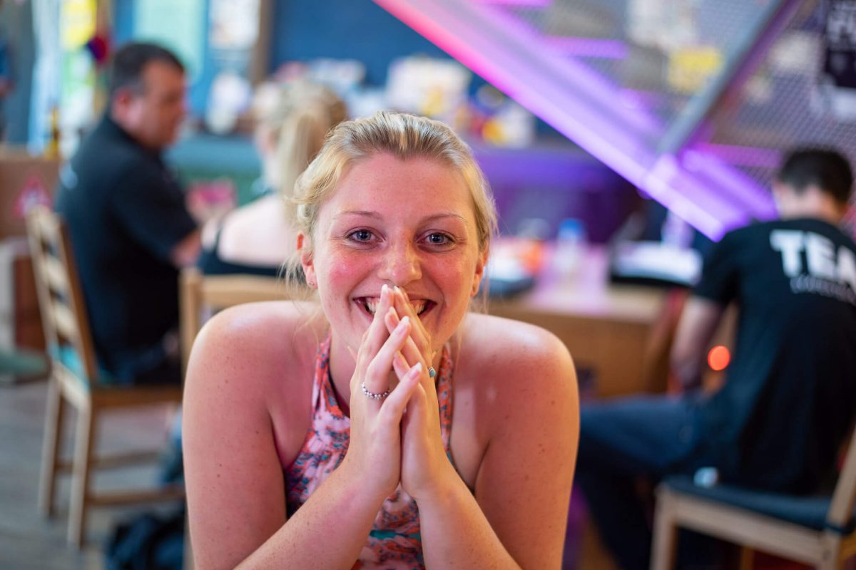 A seated performer smiles to camera with their hands clasped in front of them in a cafe