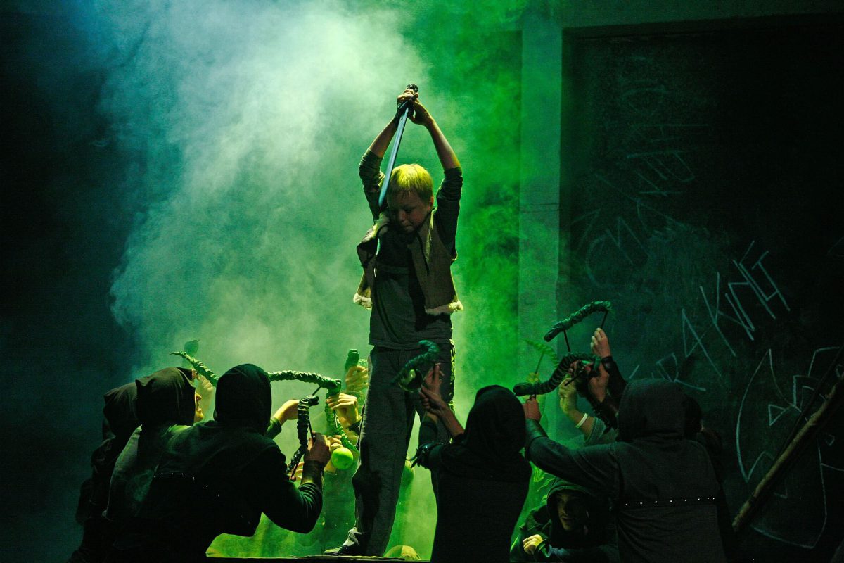 a young actor holds a sword above their head plunging it down towards the floor. They are surrounded by a group of actors.
