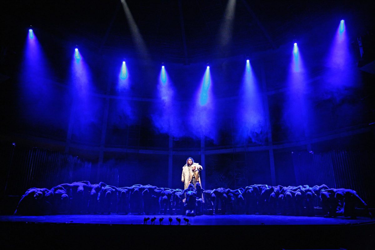 A solo performer stands in a white spotlight with dramatic blue lighting behind them