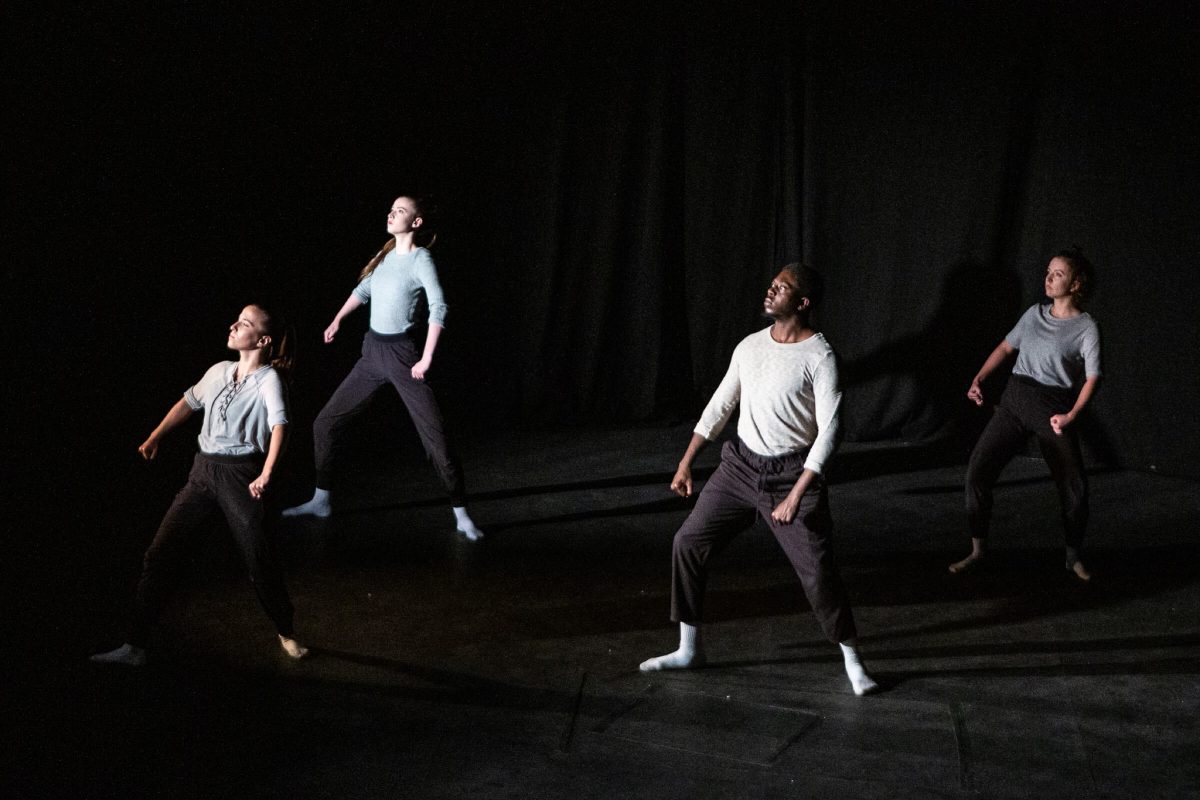 Four dancers stand in formation on stage. They are wearing grey tops and black trousers.