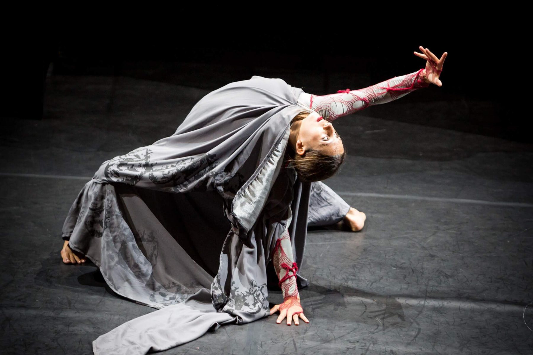 a dancer dressed in grey is crouched on the floor in a backbend with an arm outstretched behind her